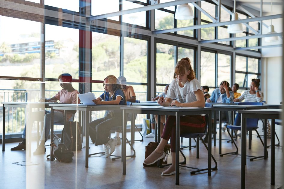 Students in classroom seated individually at single tables, with bags on floor, reading and writing on paper sheets
